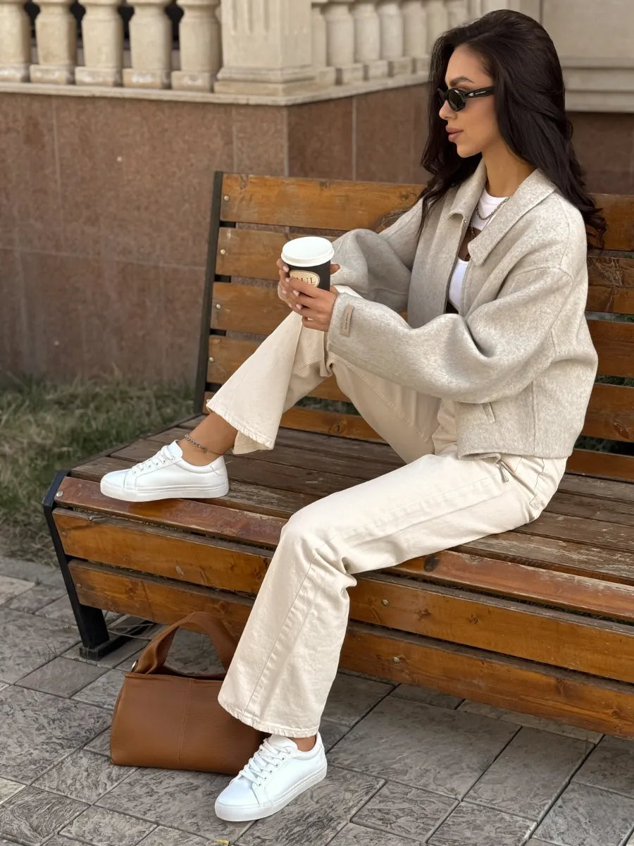 Woman sitting on a wooden bench holding a coffee cup, wearing a beige jacket and pants with white sneakers.