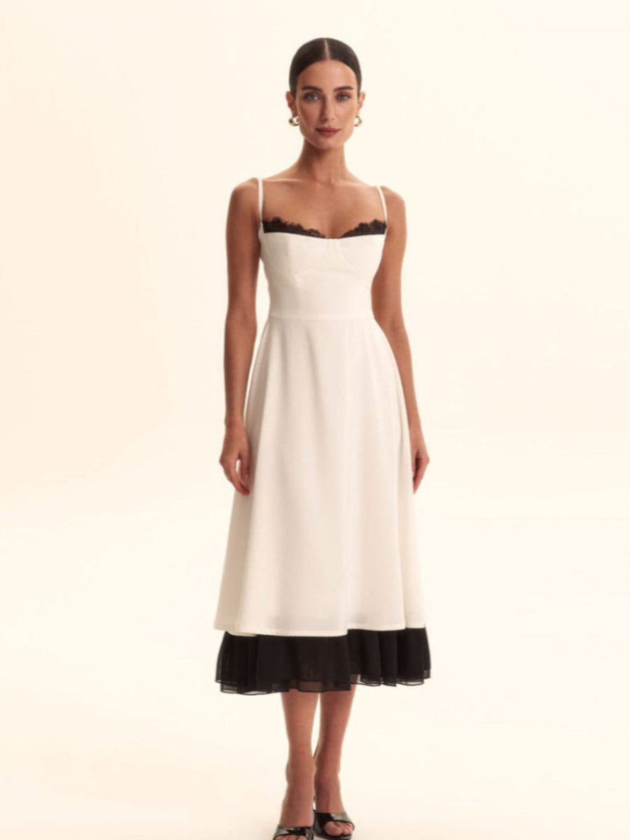A woman modeling a white midi dress with thin straps, black lace trim at the neckline, and a black ruffled hem, photographed against a plain cream studio background.