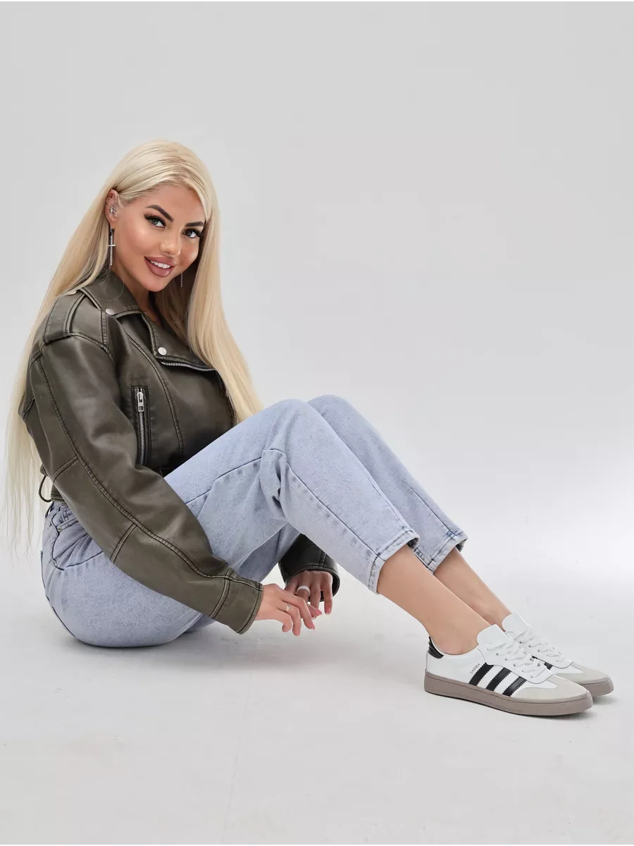 Woman wearing a brown leather jacket, light blue jeans, and white Adidas sneakers on a plain background